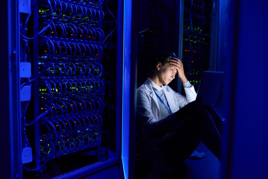 Portrait of tired young woman sitting on floor in dark server room using laptop while working with supercomputer at night